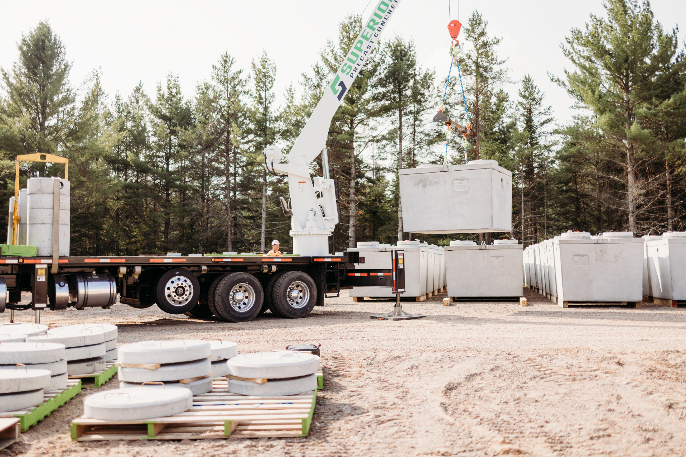 A boom truck from Superior Precast Concrete, located in Cable, Wisconsin, lifting a large precast concrete septic tank at an outdoor facility. The truck is parked beside a row of concrete tanks, with concrete lids and other products on pallets in the foreground, surrounded by pine trees.
